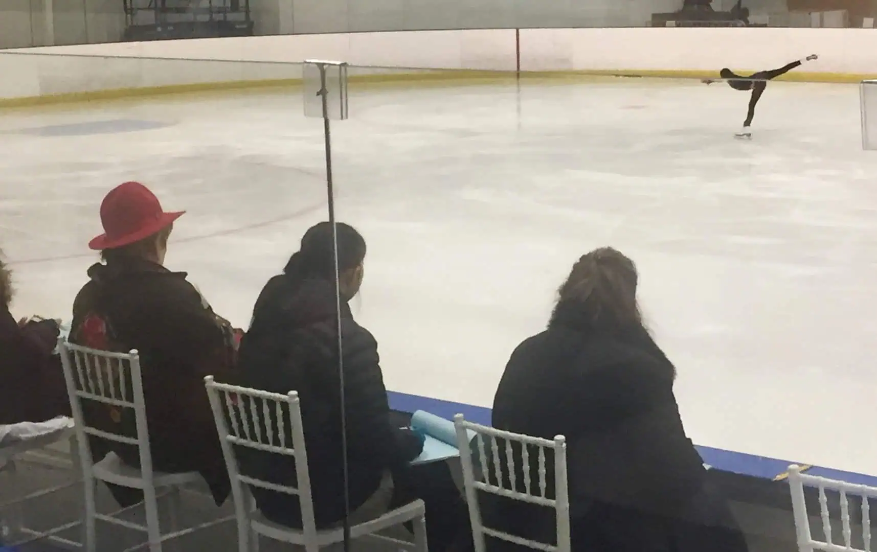 View of a female figure skater in black practice clothes doing a spiral on the ice while three adults - the judges - sit on white chairs in the hockey box watching