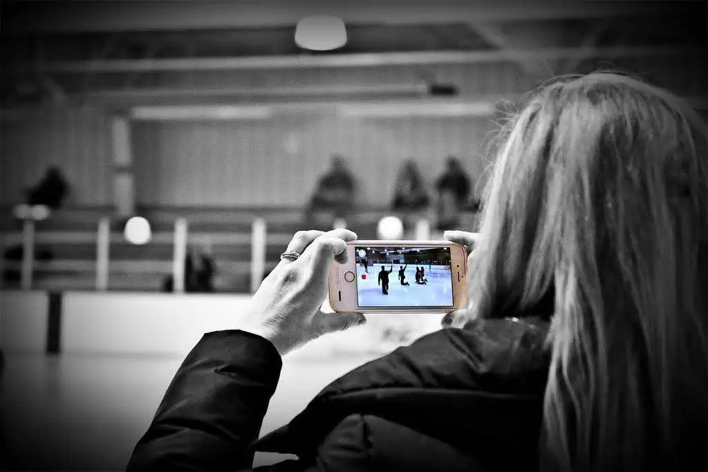 View of the back of a woman holding up a cell phone with both hands and recording a video of skaters on the ice