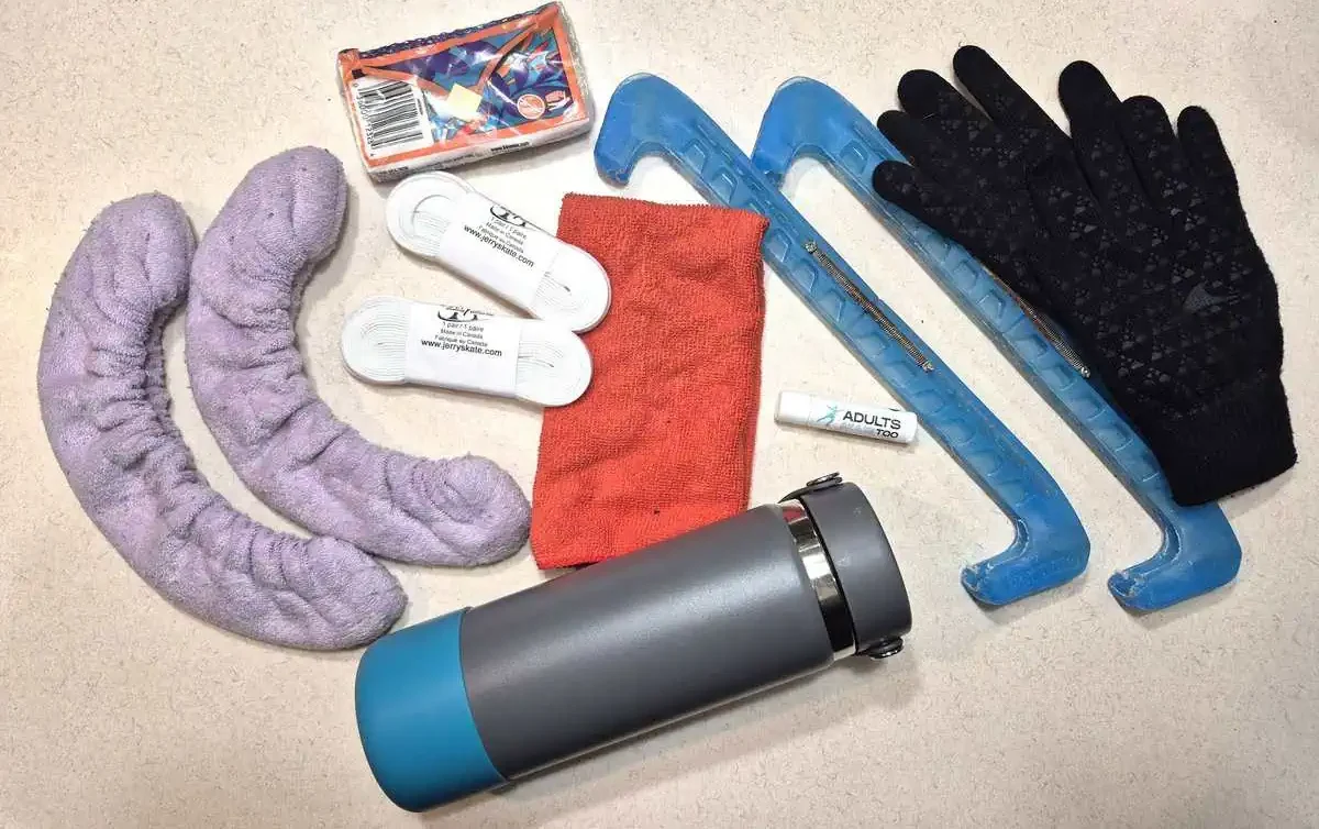 Figure skating accessories including purple soakers, two pairs of skate laces, tissues, small red absorbent towel, grey water bottle with blue base, lip balm, blue guards, and black gloves