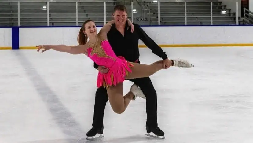 Pair skaters in a lift, the man dressed in all black and the woman in hot pink being held in front of the man