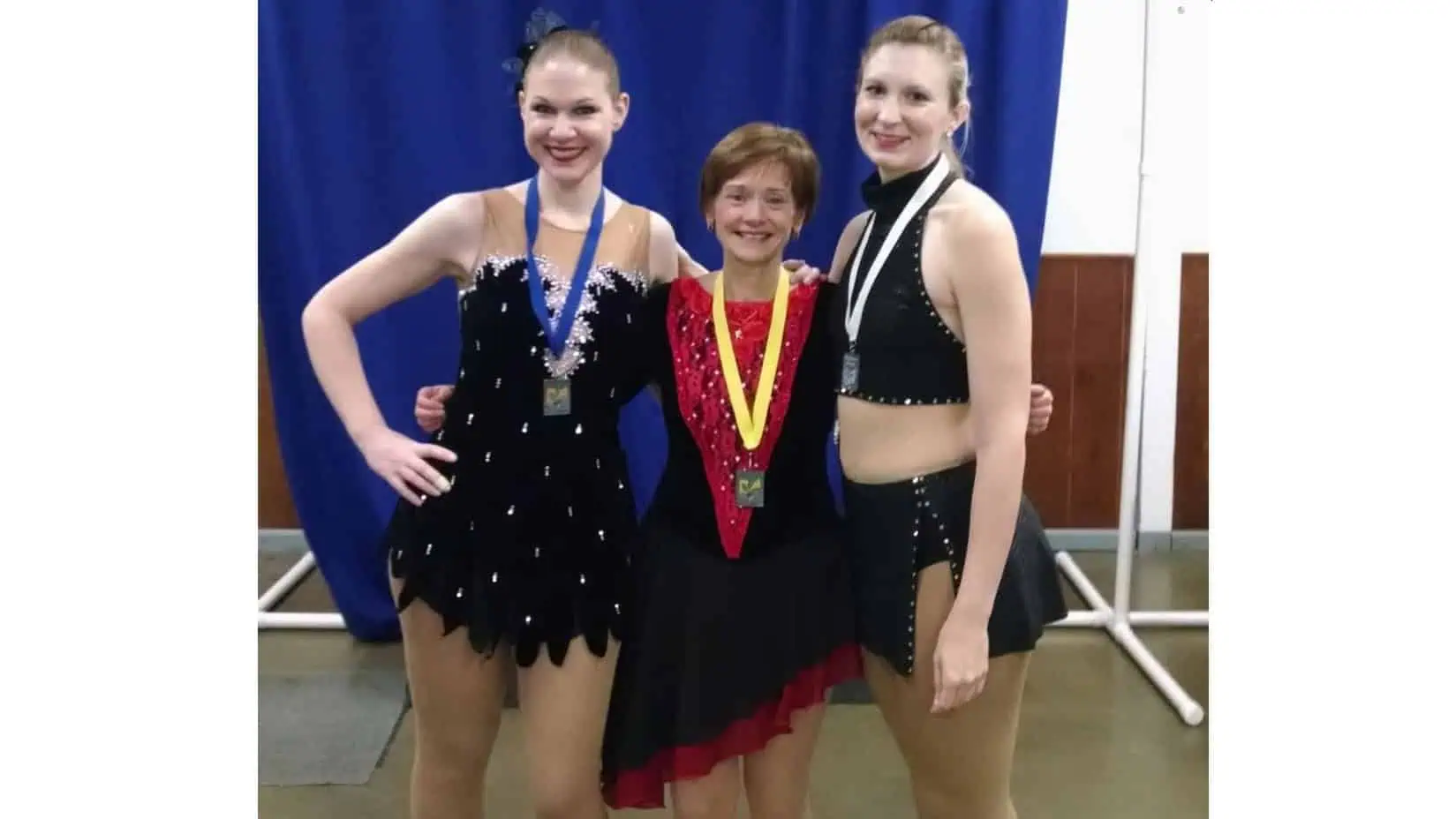 Three female adult skaters wearing their competition dresses posing with their medals around their necks: one gold, one silver and one bronze