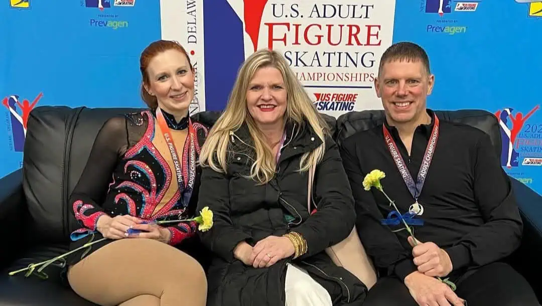 Adult pair skaters in their competition outfits sitting on a black couch on either side of their coach with the U.S. Adult Figure Skating Championships banner behind them