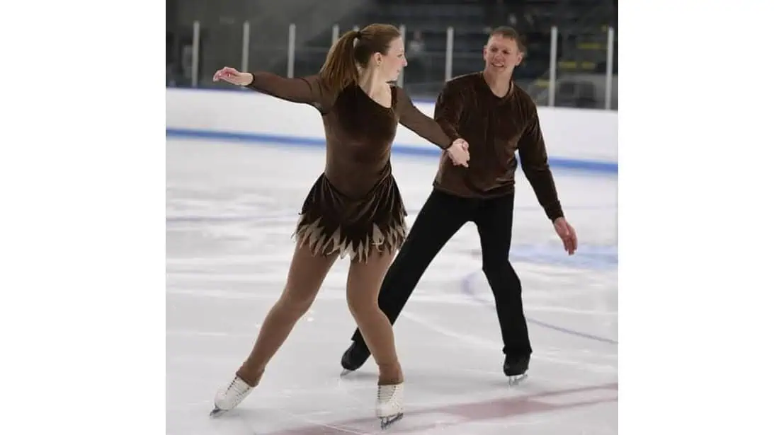 Adult pair skaters mid-program with the woman in front of and looking back at her partner and holding his hand
