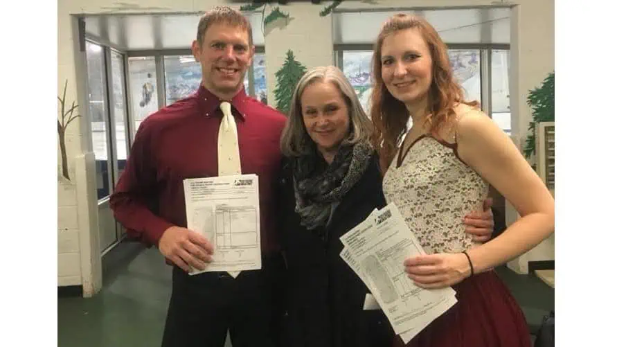 Adult ice dance partners standing on either side of their coach, holding up their test papers