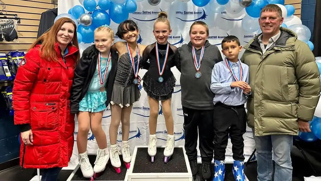 Five figure skaters, three girls and two boys, in their competition outfits and skates, posing with their medals on a podium, flanked by their coaches, one male and one female