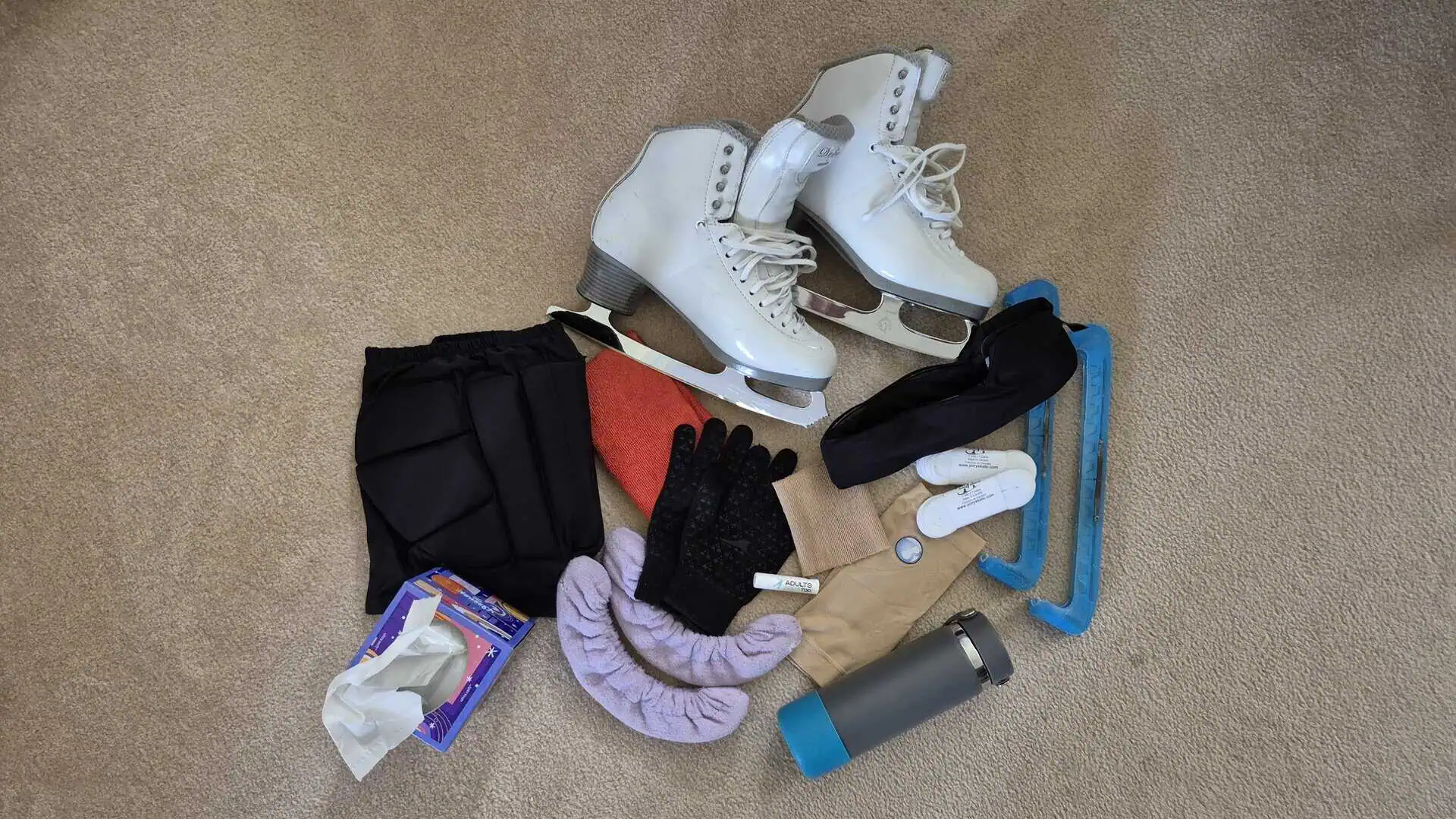 Collection of skating equipment including a box of tissues, crash pants, an absorbent towel, soakers, gloves, lip balm, Bunga pads, figure skates, head protection, skate laces, a water bottle and guards