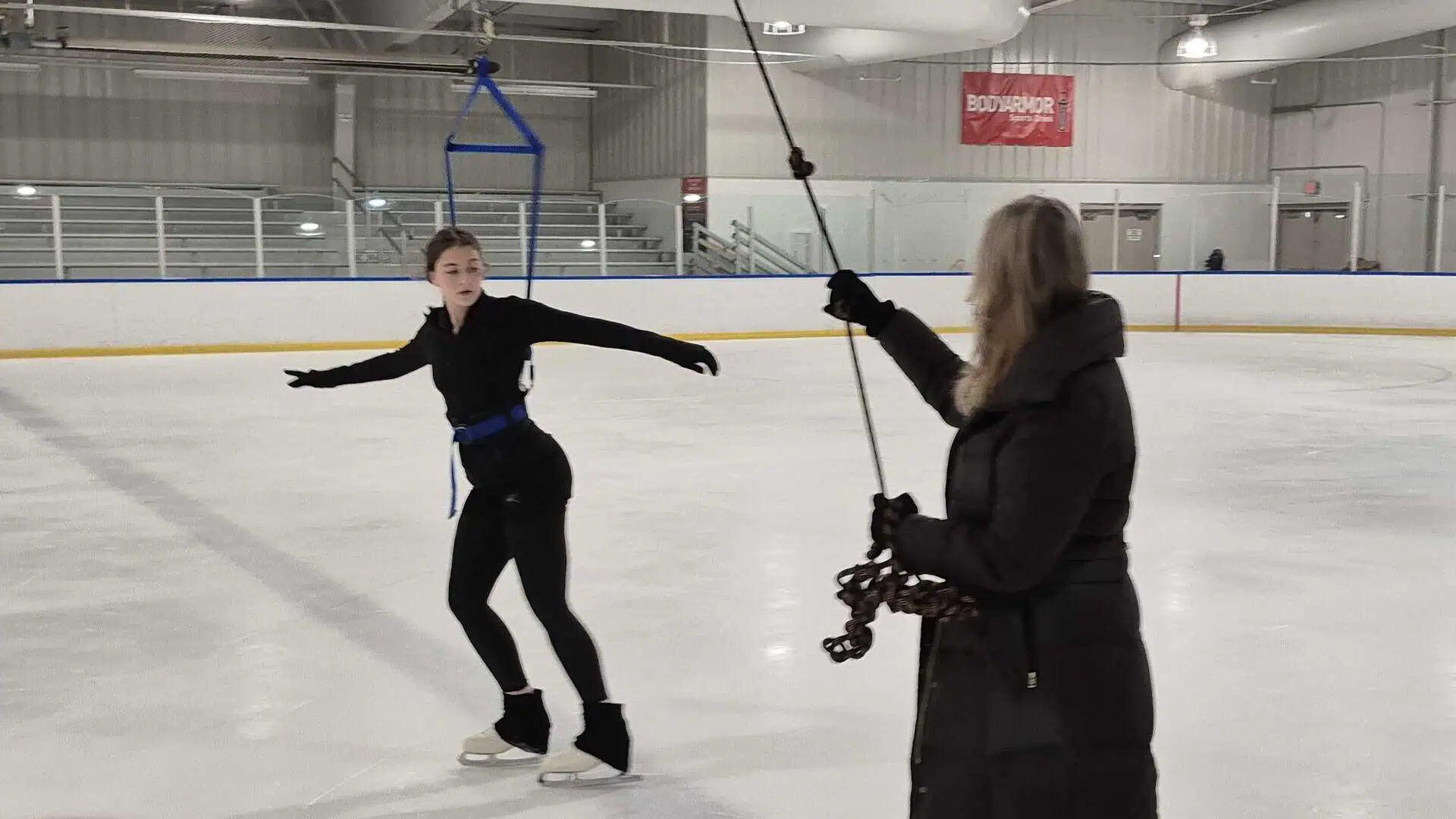 Figure skater dressed in black practice gear in the on-ice harness with her coach