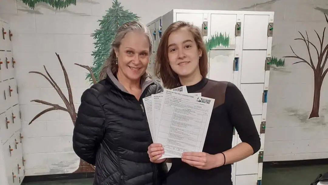 Waist-up view of a female figure skater in a three-quarter-sleeved black skating dress standing next to her coach who is wearing a black winter coat, holding up three fanned-out test papers in front of them, with both people smiling