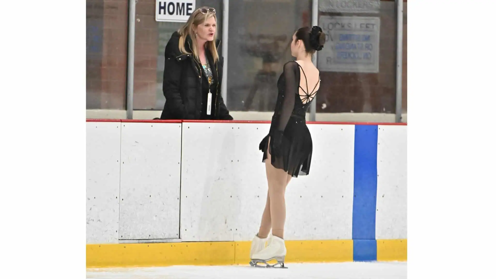 Figure skater in black dress standing at the boards by the hockey box getting feedback from her coach