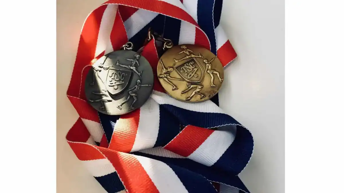 A silver and a gold figure skating medal with skaters shown performing various skating elements, lying on red, white, and blue striped ribbon