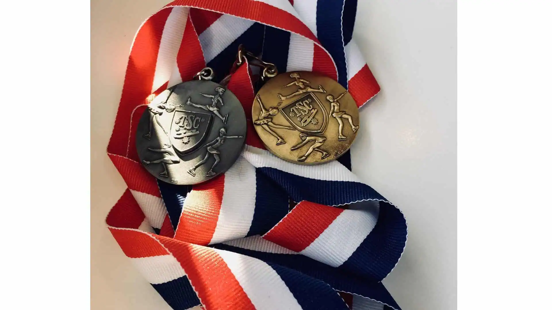 A silver and a gold figure skating medal with skaters shown performing various skating elements, lying on red, white, and blue striped ribbon