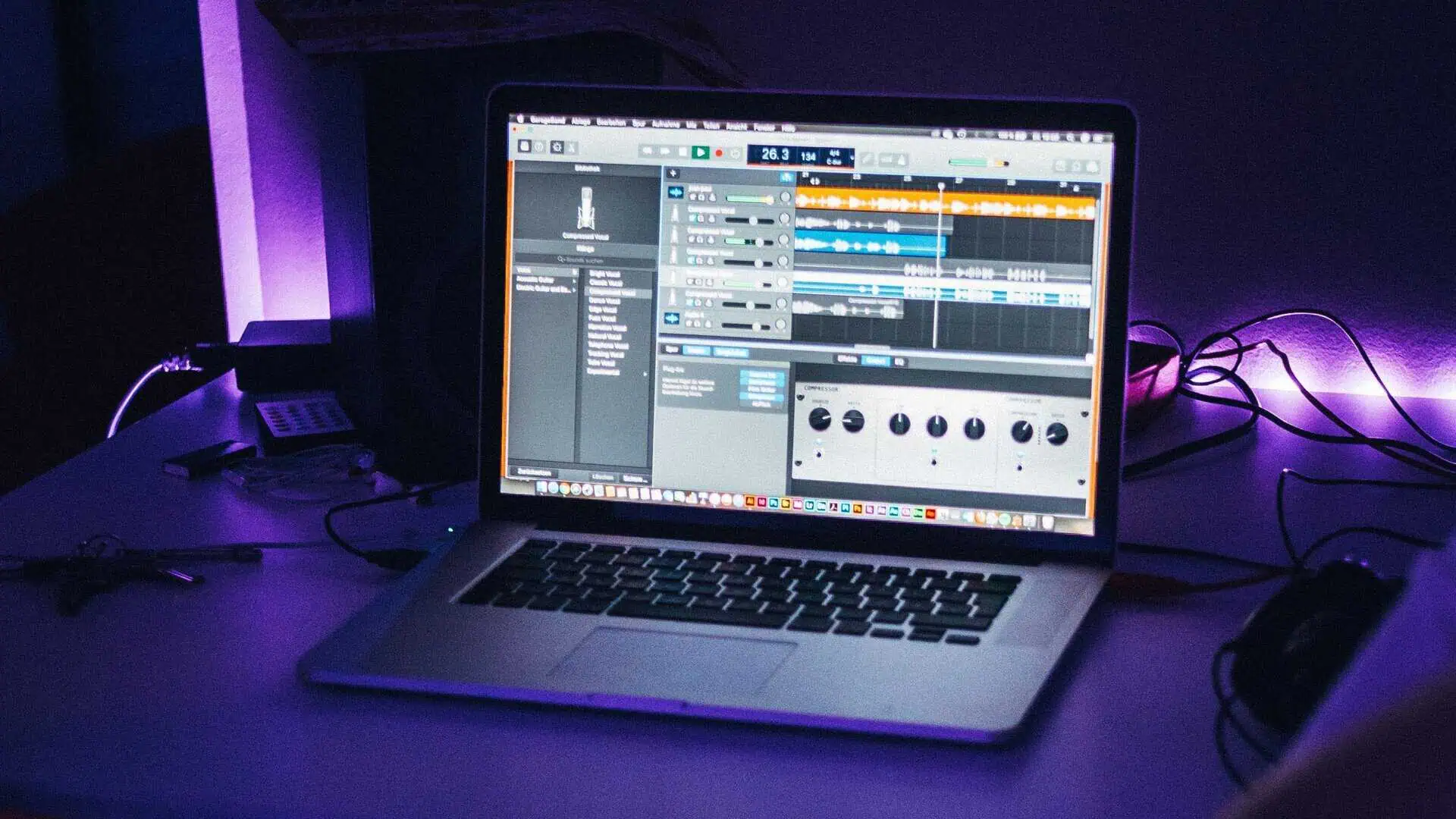 Laptop sitting on a table and showing music being cut on the screen, with various cords around it and purple lighting in the background Sound waves represented digitally as bars ranging in color from green to yellow to orange to red going from bottom to top