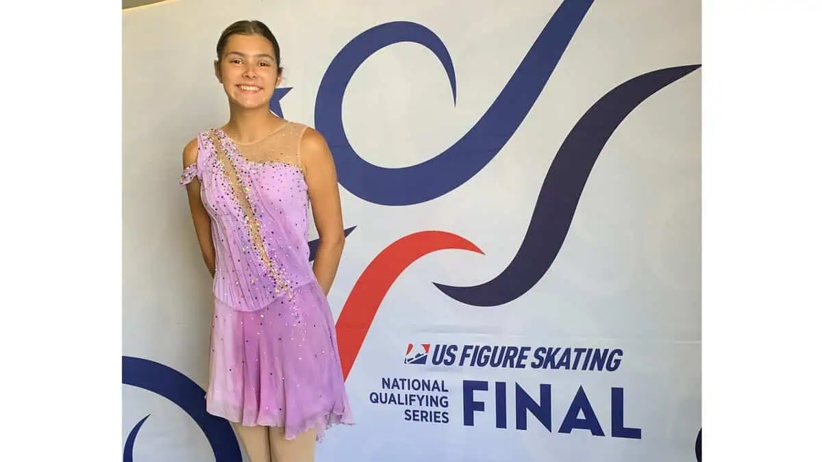 Teen figure skater in a lavender skating dress standing in front of a U.S. Figure Skating National Qualifying Series Final banner