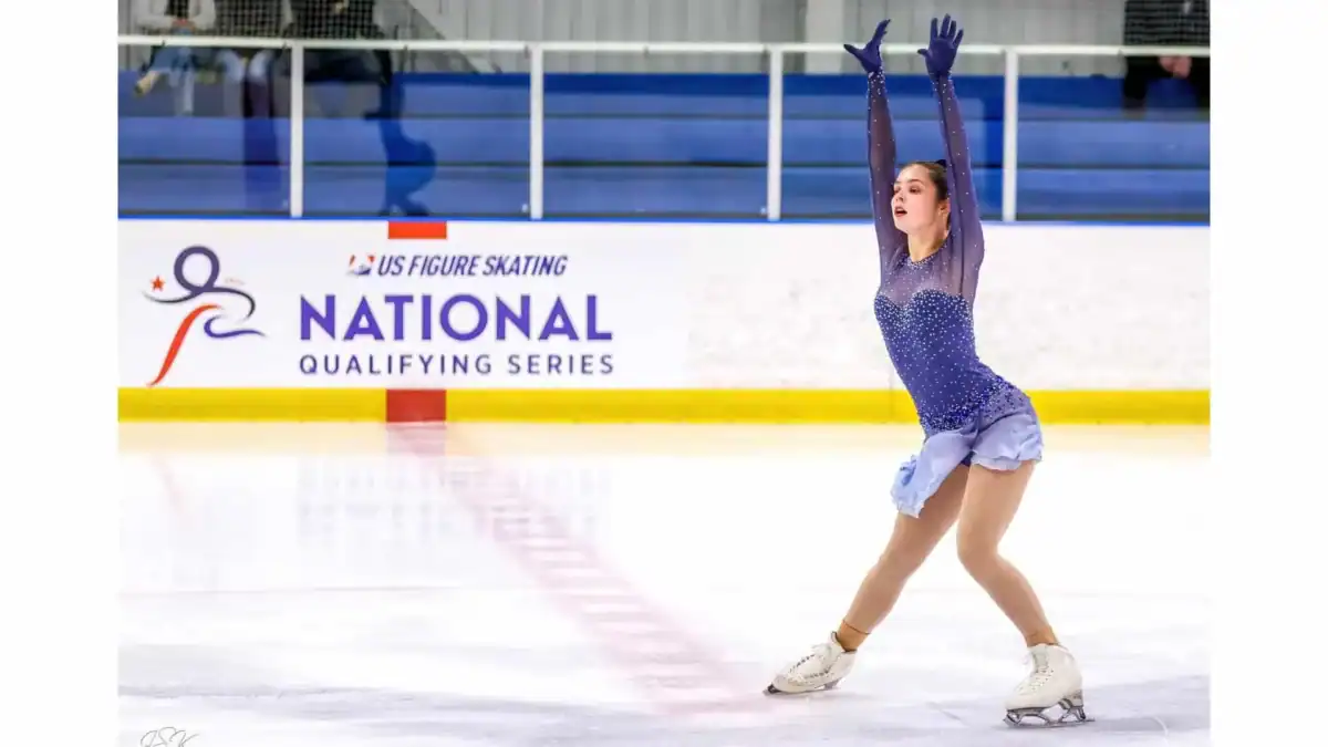 Figure skater in a blue dress performing at a National Qualifying Series competition as she skates past the U.S. Figure Skating National Qualifying Series sign on the side of the rink