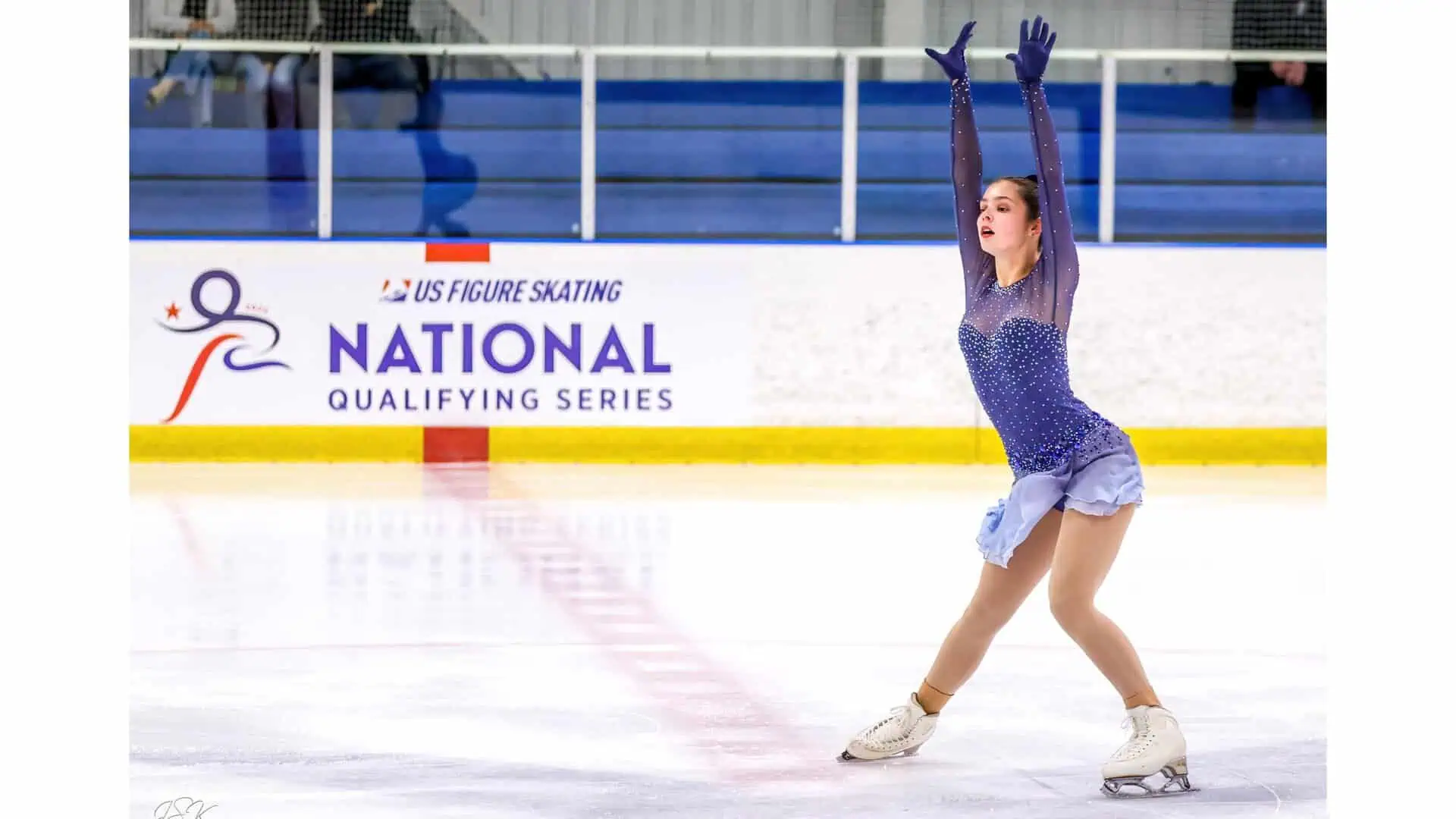 Figure skater in a blue dress performing at a National Qualifying Series competition as she skates past the U.S. Figure Skating National Qualifying Series sign on the side of the rink