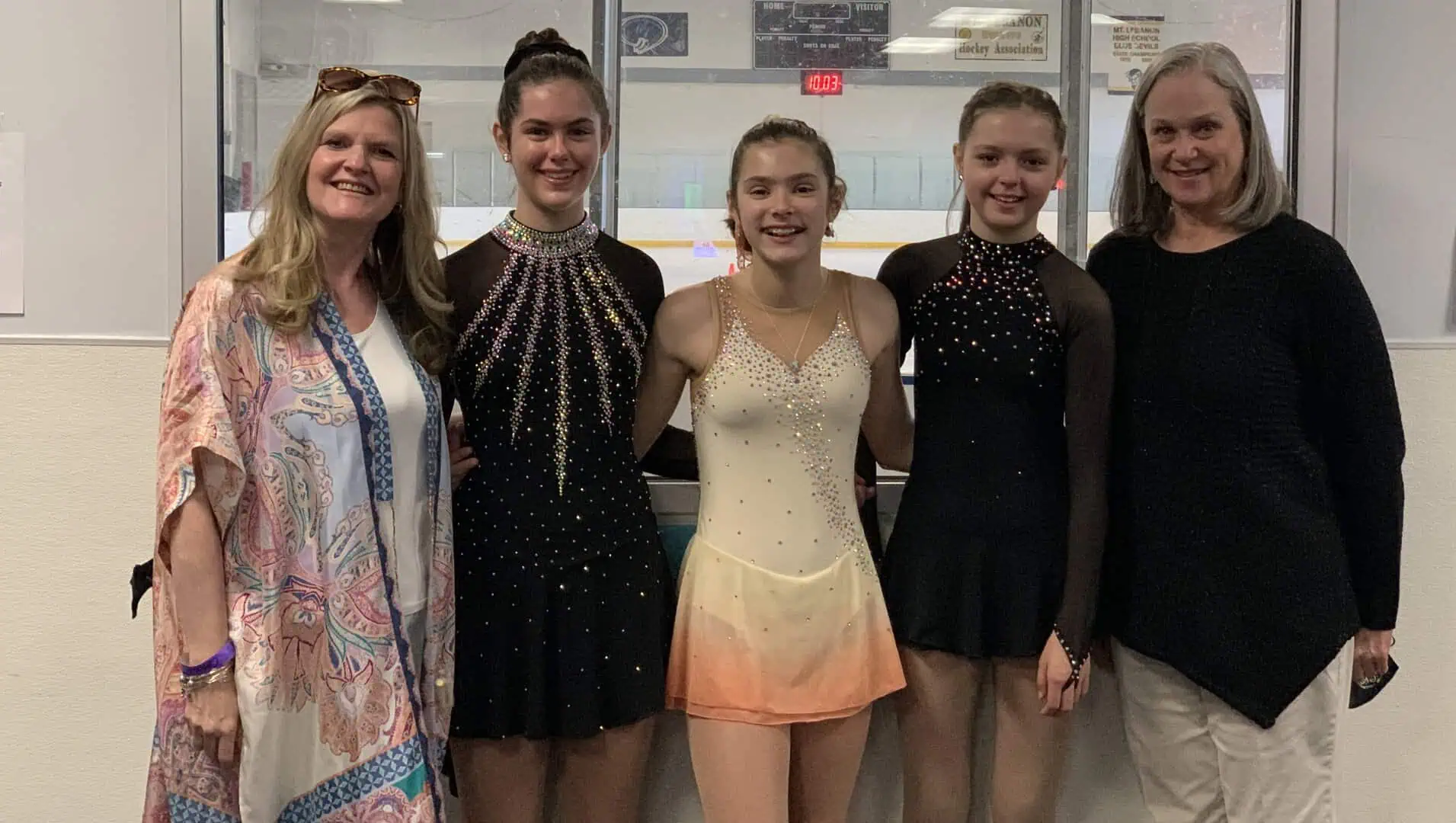 Three girls in skating dresses, two in black and one in peach, standing side by side, flanked by their two coaches, with the sheet of ice in the background