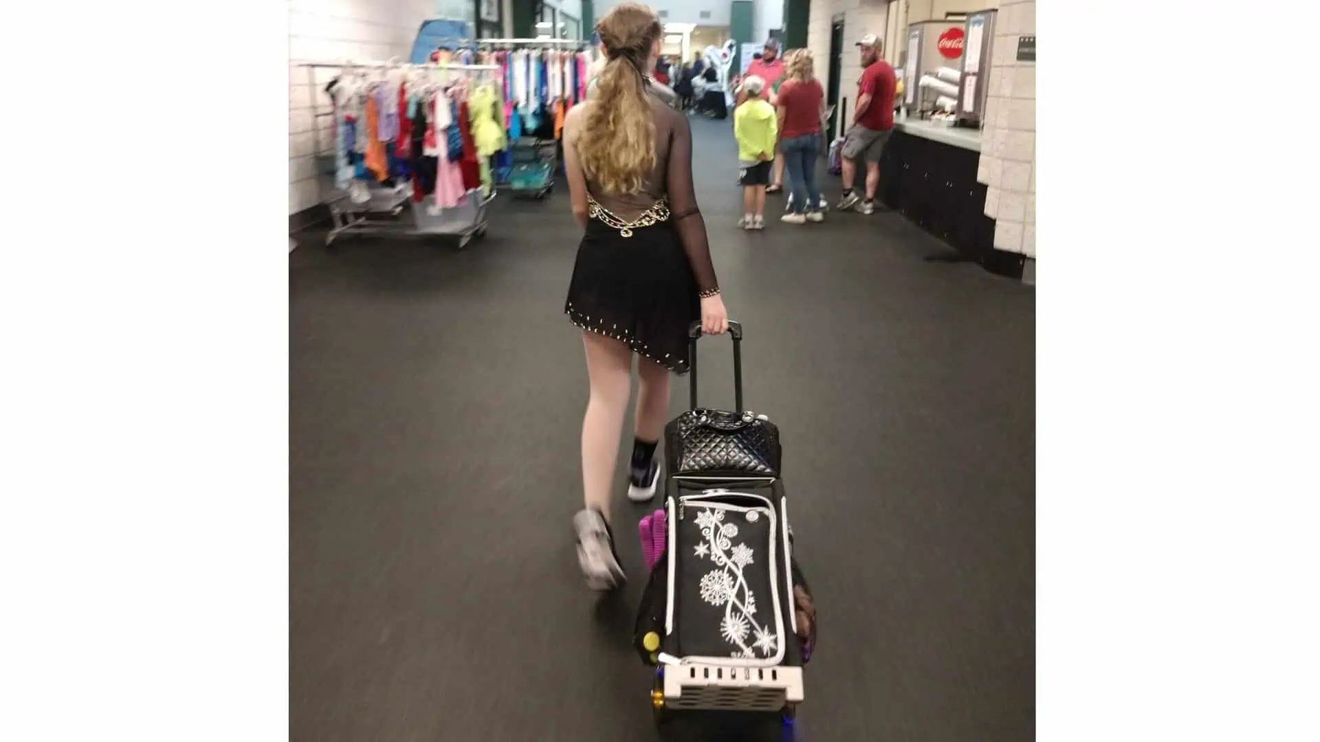Rear view of a figure skater in a black dress and sneakers arriving at a competition, pulling her skating bag behind her and looking for a place to set up and prepare for her event